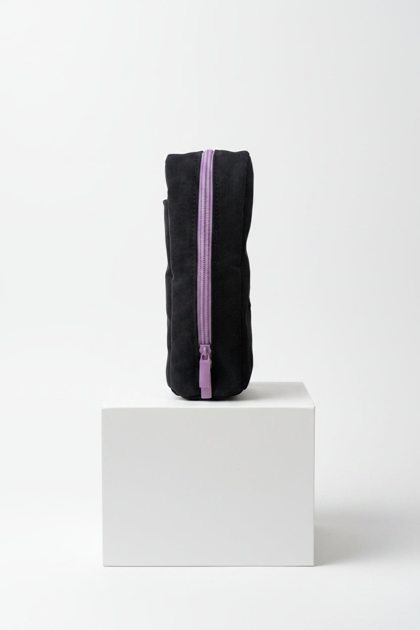 A black fabric pencil case with a prominent purple zipper, held by a hand on a white tabletop with a blurred indoor background.