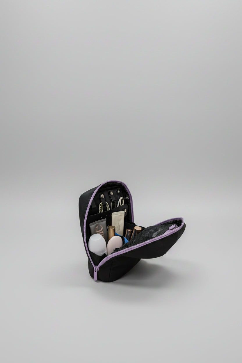 A black makeup bag with purple trim, shown open on a white table with various cosmetics inside. The background is a blurred indoor room.