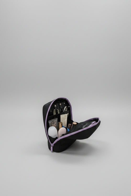 A black makeup bag with purple trim, shown open on a white table with various cosmetics inside. The background is a blurred indoor room.